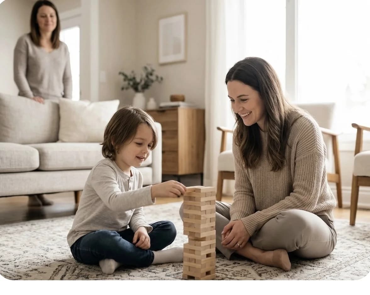 A nanny building a block tower with a young child on the living room floor while the mother looks on